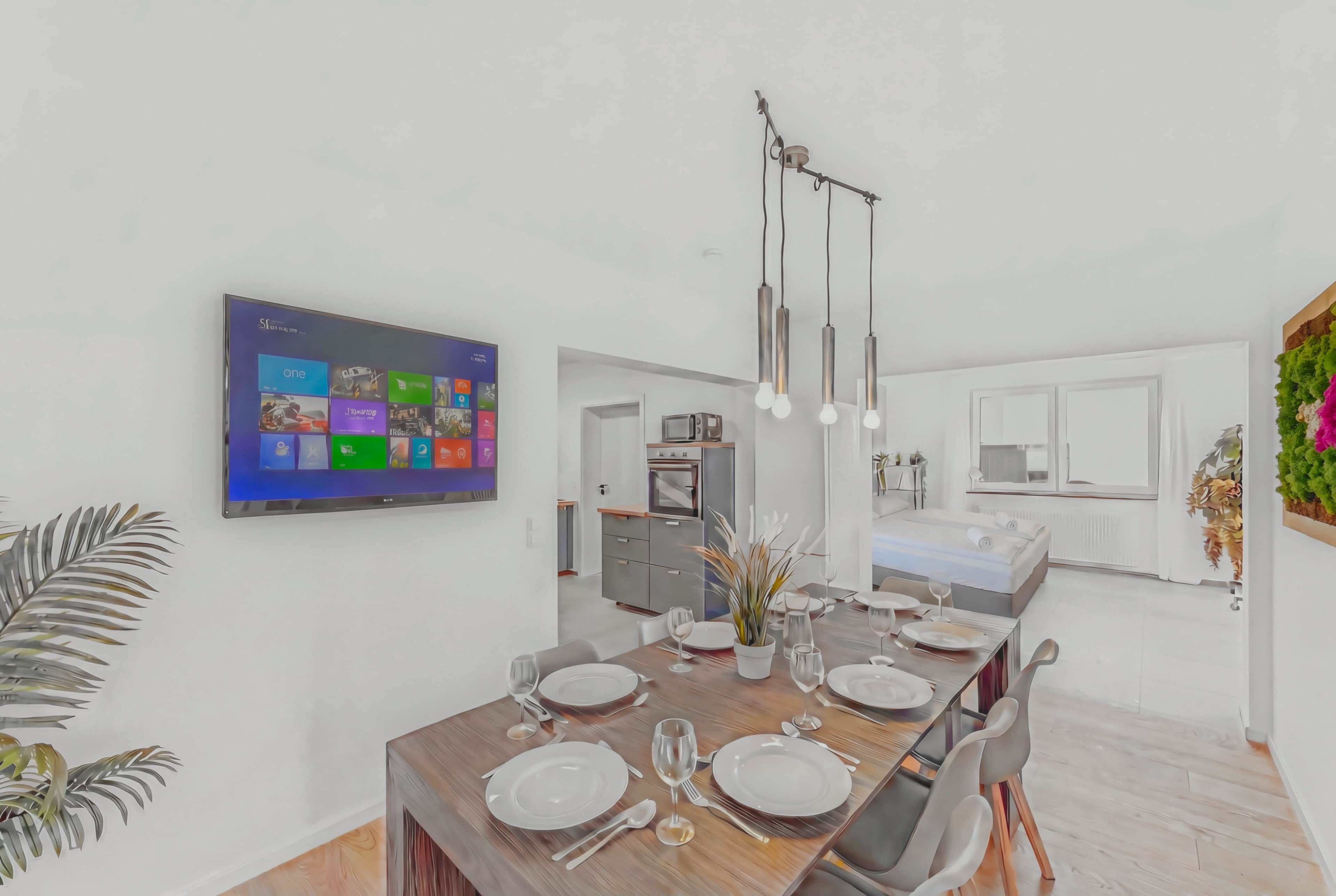 Een moderne eethoek met een lange tafel gedekt voor een maaltijd, een flatscreen televisie aan de muur en een keuken op de achtergrond.
