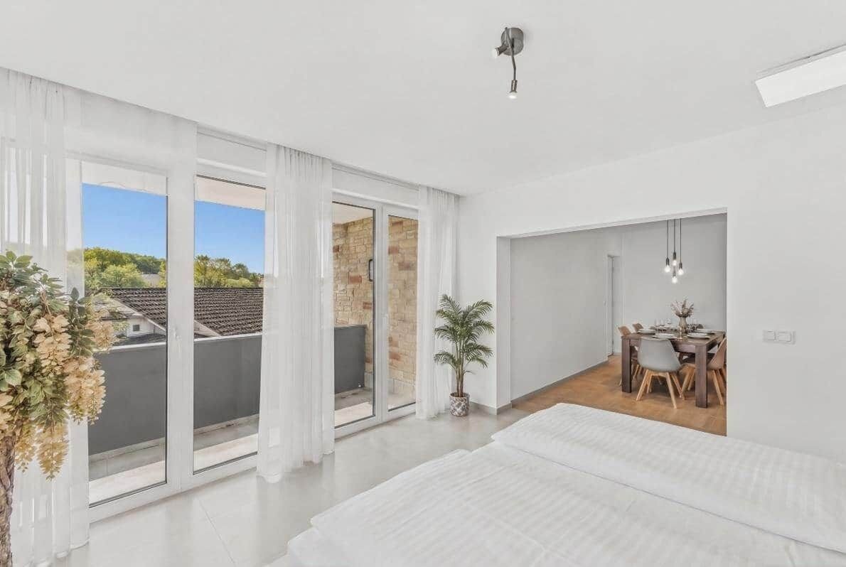 Ein modernes Schlafzimmer mit einem großen Bett, einem Essbereich mit Tisch und Stühlen sowie ein Blick durch eine Glastür auf einen Außenbalkon.