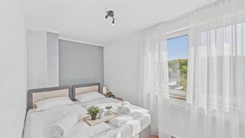 Ein modernes Schlafzimmer mit einem Bett, Nachttischen, einem Fenster mit leichtem Vorhang und Blick auf Grünflächen.