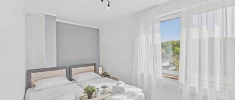A modern bedroom with a bed, bedside tables, a window with sheer curtains, and a view of greenery.