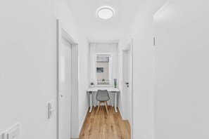 Un pasillo estrecho con un escritorio y una silla blancos, una lámpara en el techo y una puerta que da a otra habitación.