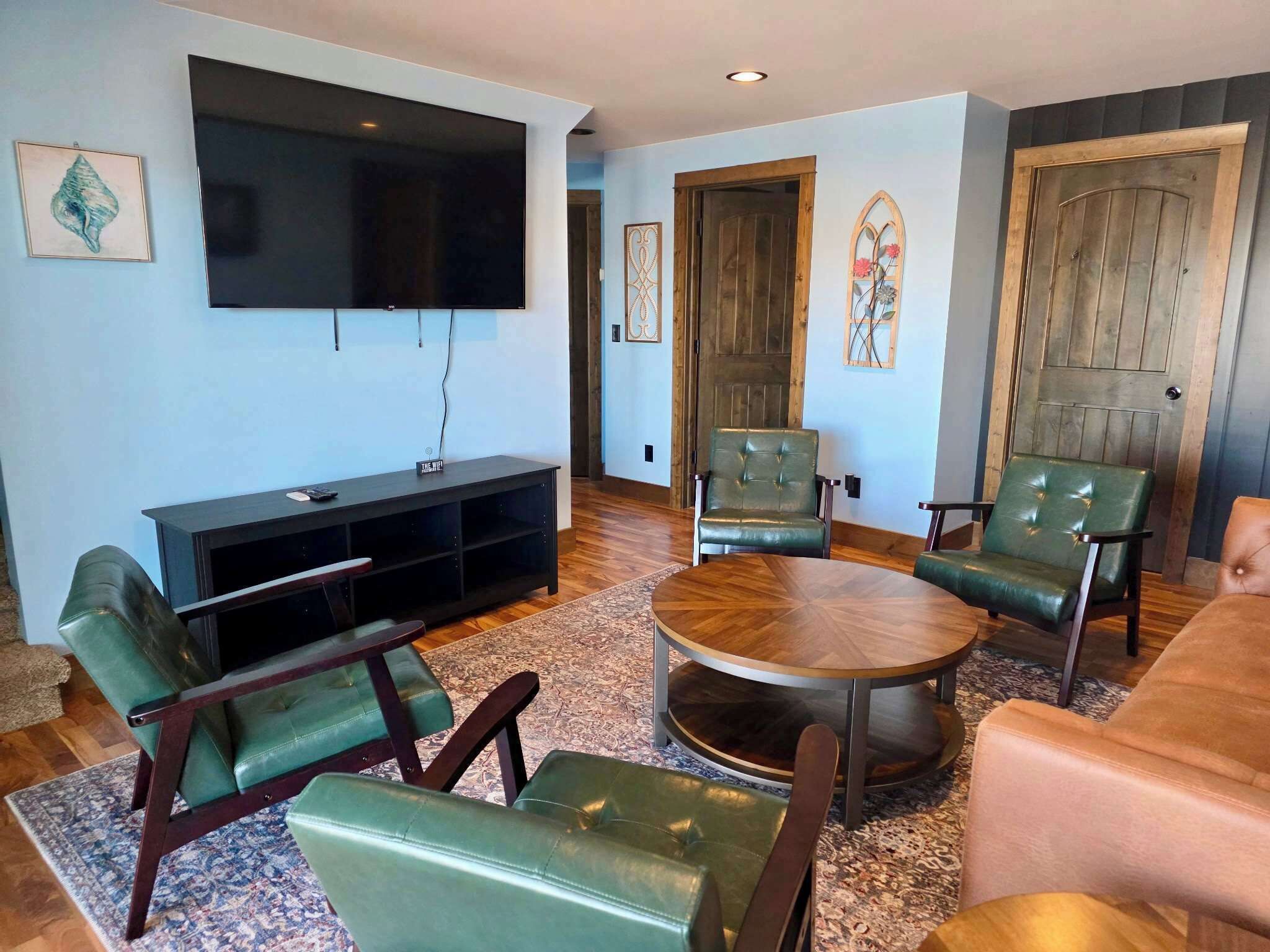 A living room with a flat-screen TV, green leather chairs, a round coffee table, and a wooden door.