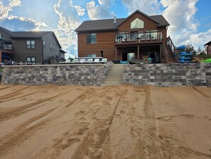 A two-story wooden house with a balcony, stone walls, and a sandy area in front.