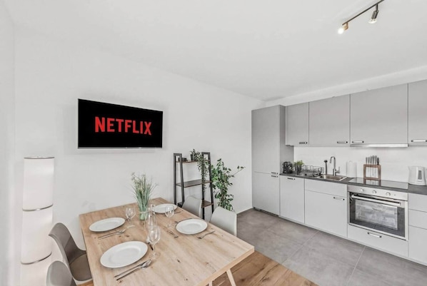 Una cocina moderna con armarios blancos, un comedor con mesa de madera dispuesta para cuatro personas, y un televisor de pared mostrando 'NETFLIX'.
