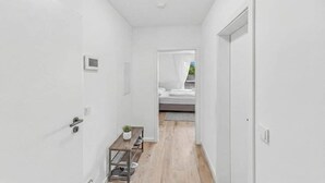 Un pasillo estrecho con paredes blancas, piso de madera, una mesita con una planta y una cama al fondo.