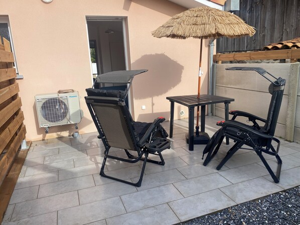 Une terrasse extérieure avec une table et deux chaises, un parasol en chaume et un appareil de climatisation.
