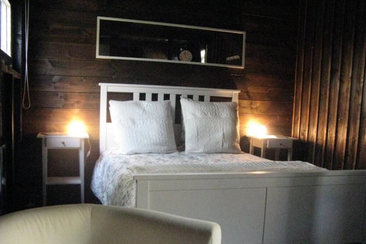 Una cama con ropa de cama blanca, dos mesitas de noche con lámparas, una silla y una pared de madera con un espejo sobre la cama.