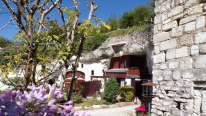 Un edificio de piedra con un exterior rústico, un jardín con arbustos y un árbol, y un cielo despejado.