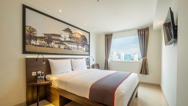 Quarto de hotel com cama, mesa de cabeceira com garrafas de água, quadro na parede, janela com cortinas e televisão fixada na parede.