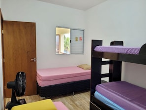 Une chambre de dortoir avec des lits superposés, un lit simple, une porte en bois, une fenêtre donnant sur un paysage agréable et un ventilateur sur pied.