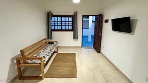 Een kamer met een houten bankje, een vloerkleed, een aan de muur bevestigde televisie en een raam met gordijnen.