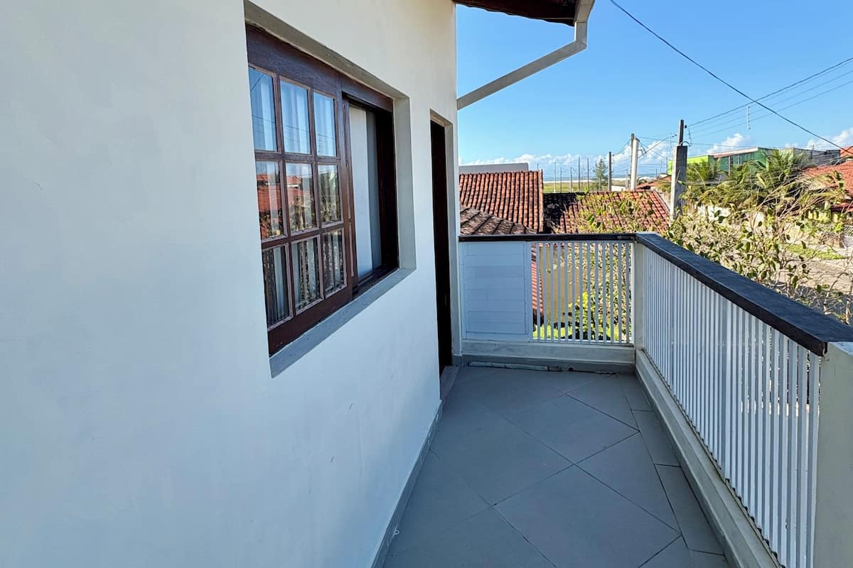 A balcony with a tiled floor, a window with a wooden frame, and a view of a residential area with houses and a clear sky.