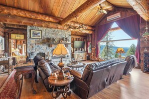 Ein Blockhaushaus-Interieur mit Kamin aus Stein, Ledermöbeln, einem Fernseher und Blick ins Freie.