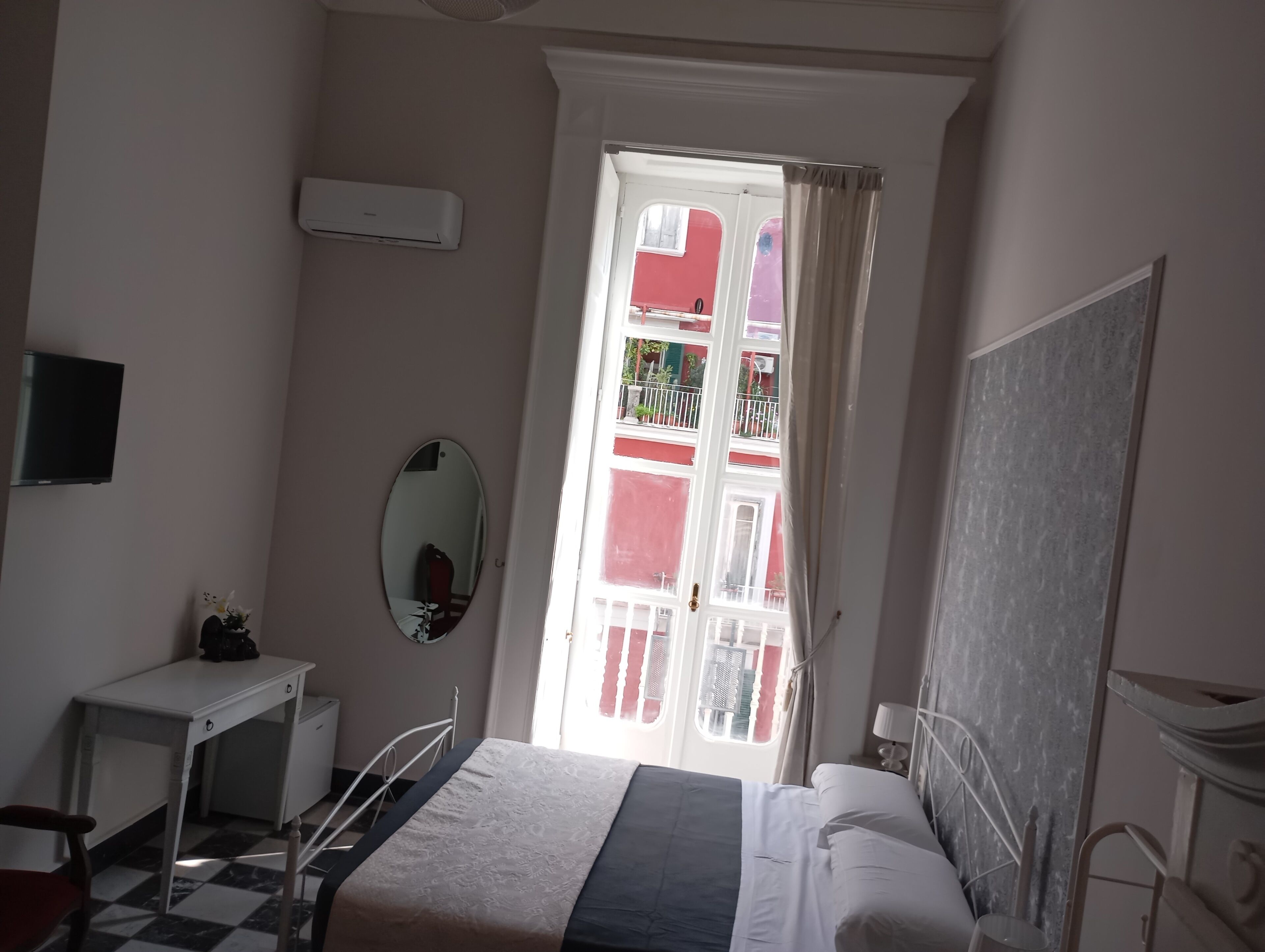 Een slaapkamer met een bed, een bureau, een spiegel en een raam met uitzicht op een balkon.