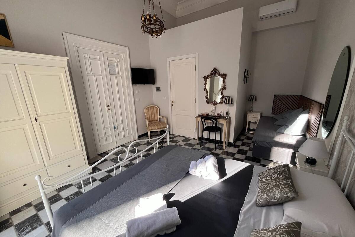 A hotel room with a checkered floor, a bed with pillows, a desk with a chair, a wardrobe, and a mirror.