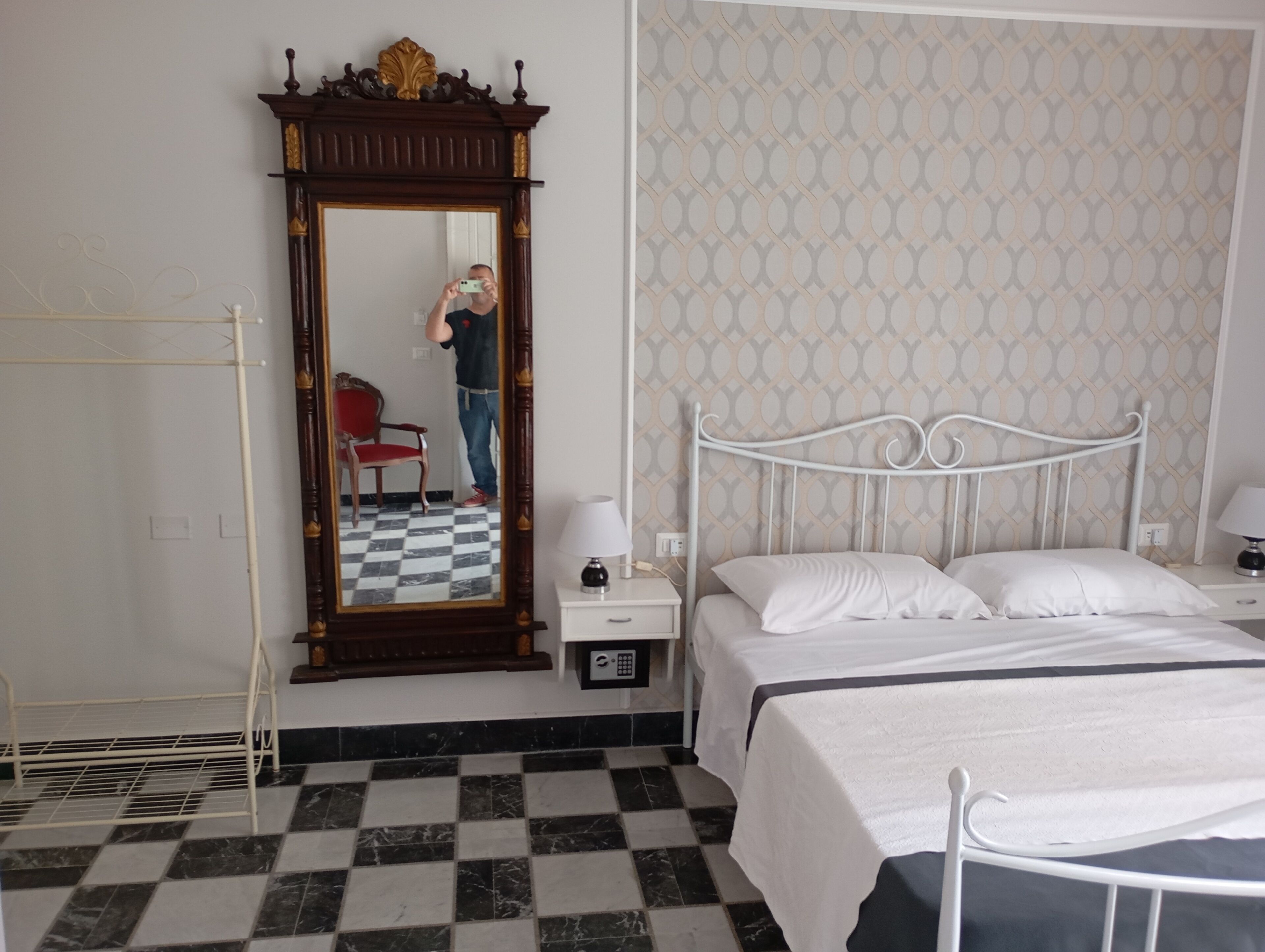A hotel room with a checkered floor, a bed with white linens, a nightstand with a lamp, a mirror with a wooden frame, and a patterned wallpaper.