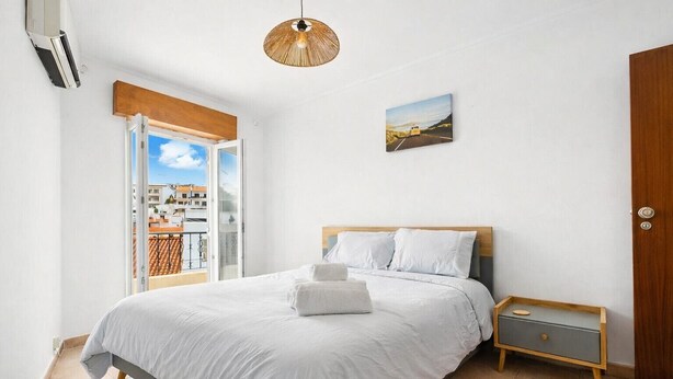 A bedroom with a bed, a nightstand, a wooden door, a picture on the wall, and a window with a view of buildings. - 2-bedr. apartment Palhinhas Historic Center 5 min. away (Albufeira)