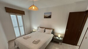 A bedroom with a bed, two bedside tables, a hanging light fixture, a door, and a window with blinds. - 2-bedr. apartment Palhinhas Historic Center 5 min. away (Albufeira)