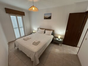 Ein Schlafzimmer mit einem Bett, zwei Nachttischen, einer Hängeleuchte, einer Tür und einem Fenster mit Jalousien.