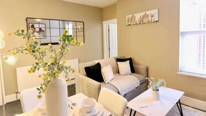 Ein Wohnzimmer mit einer Couch, einem Esstisch mit Stühlen, einer Vase mit Blumen und einem Bild an der Wand.