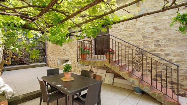 Un espace extérieur entouré de murs en pierre, avec une table et des chaises, un escalier doté d’une rampe en métal et une plante en pot.