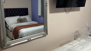 Habitación de hotel con un espejo grande y ornamentado, un televisor de pantalla plana, un tocador blanco con un organizador transparente y una cama con ropa de cama blanca y una manta marrón.