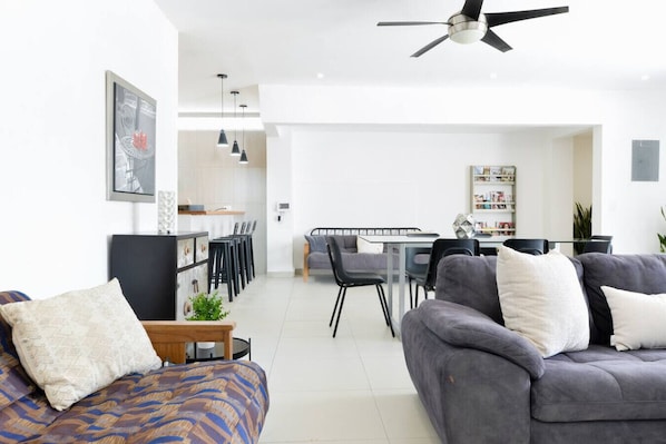A modern living room with a grey sofa, a patterned sofa, a dining area with a glass table, and a ceiling fan.