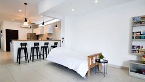 Une pièce moderne et bien éclairée, comprenant un lit, une petite table avec une plante et un coin cuisine avec des tabourets de bar.