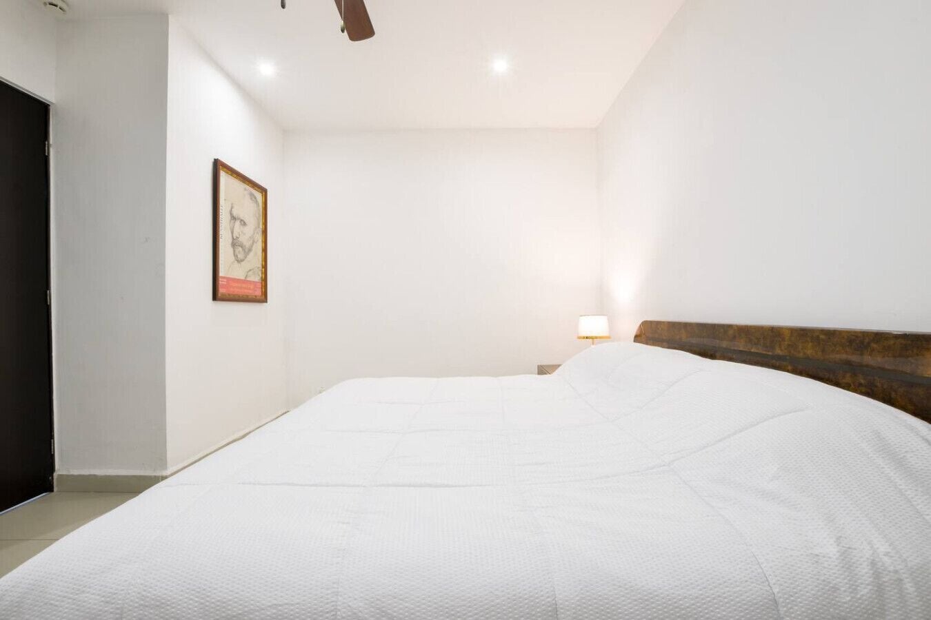 A bedroom with a large bed, a nightstand with a lamp, a framed picture on the wall, and a ceiling fan.