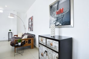 A modern hotel room with a bed, a side table with a plant, a black dresser with decorative items, and wall art.