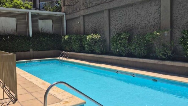 A swimming pool with a metal ladder, surrounded by a brick wall and a building with multiple windows.