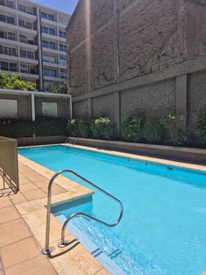 Una piscina con escalera metálica, rodeada por un muro de ladrillo y un edificio con múltiples ventanas.