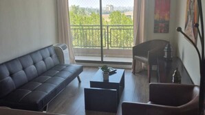 A modern living room with a sofa, coffee table, and chair, a balcony with a view of greenery and buildings, and a painting on the wall.