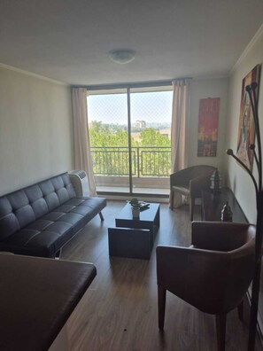 A modern living room with a sofa, coffee table, and chair, a balcony with a view of greenery and buildings, and a painting on the wall. - Modern Apartment - Steps away from Movistar Arena and Hipódromo (Santiago)