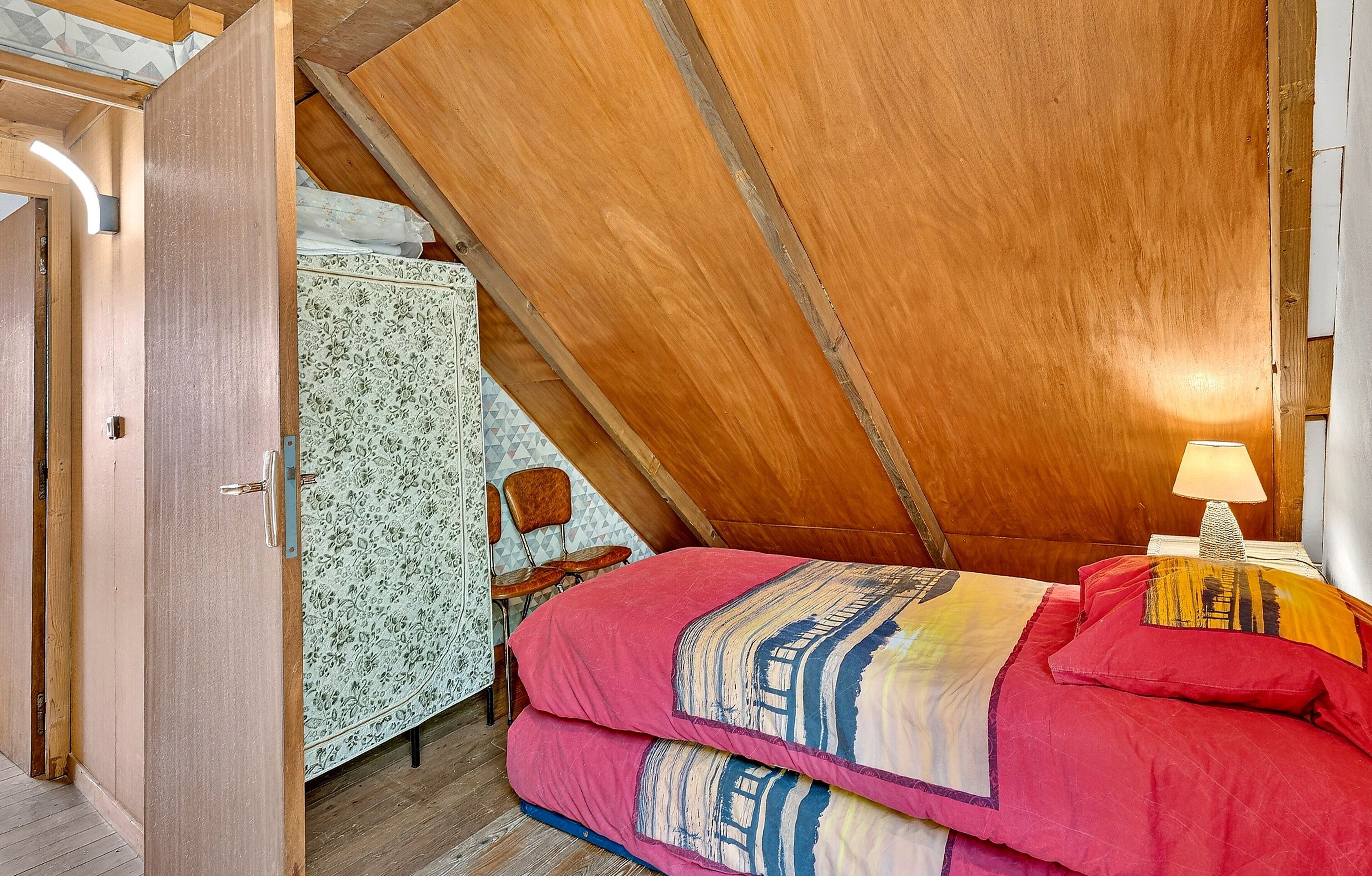 Un dormitorio con techo inclinado, una cama con colcha estampada, una mesita de noche con lámpara y una silla.
