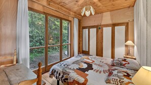 A wooden bedroom with large windows, a bed with a floral bedspread, a hanging light fixture, and a bedside table with a lamp. - Chalet with Mountain Views, Private Garden and Balcony (Seyne)