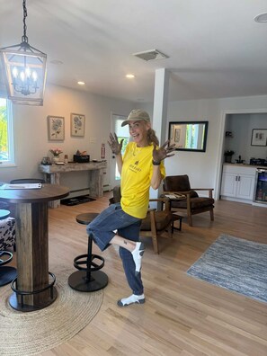 A person in a yellow shirt dancing in a room with a wooden floor, a round table, and a bar area in the background.