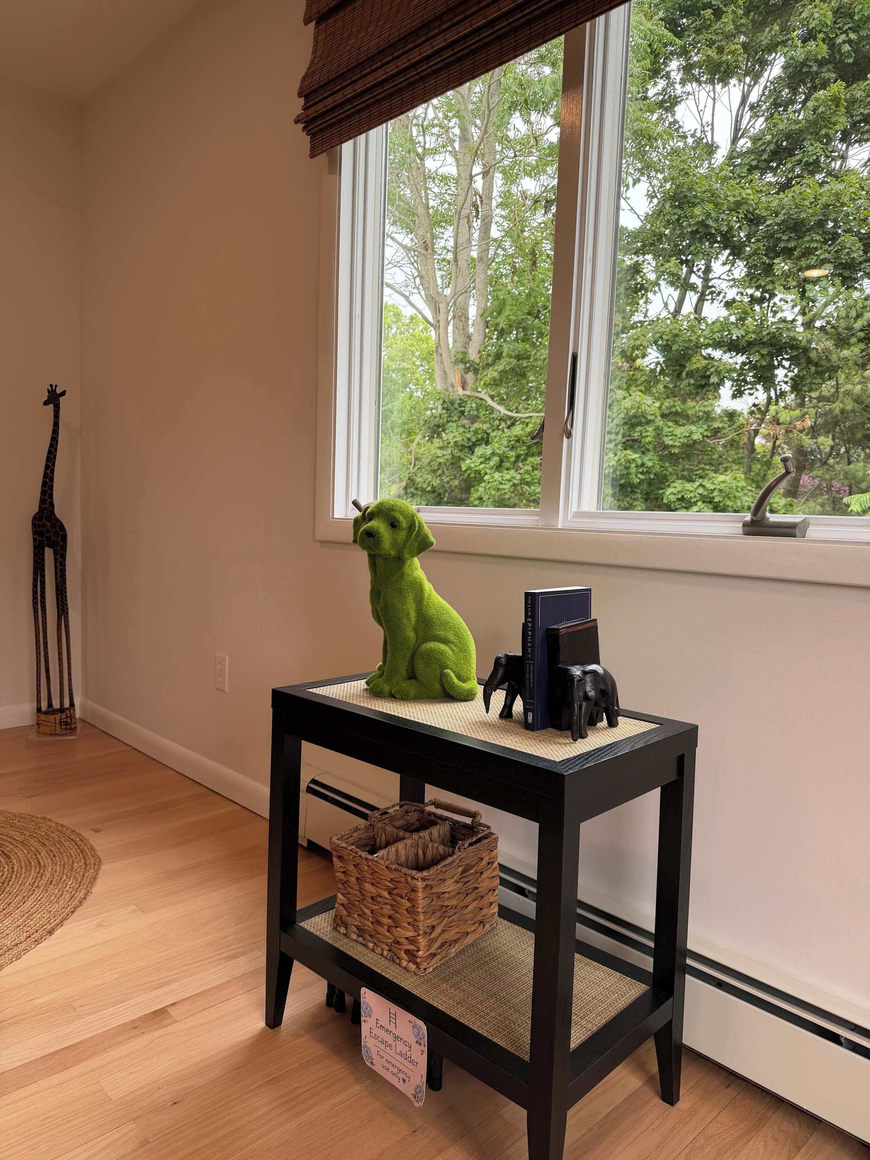 Une pièce avec une fenêtre, une petite table sur laquelle se trouve un panier, une décoration représentant une girafe et un animal en peluche.