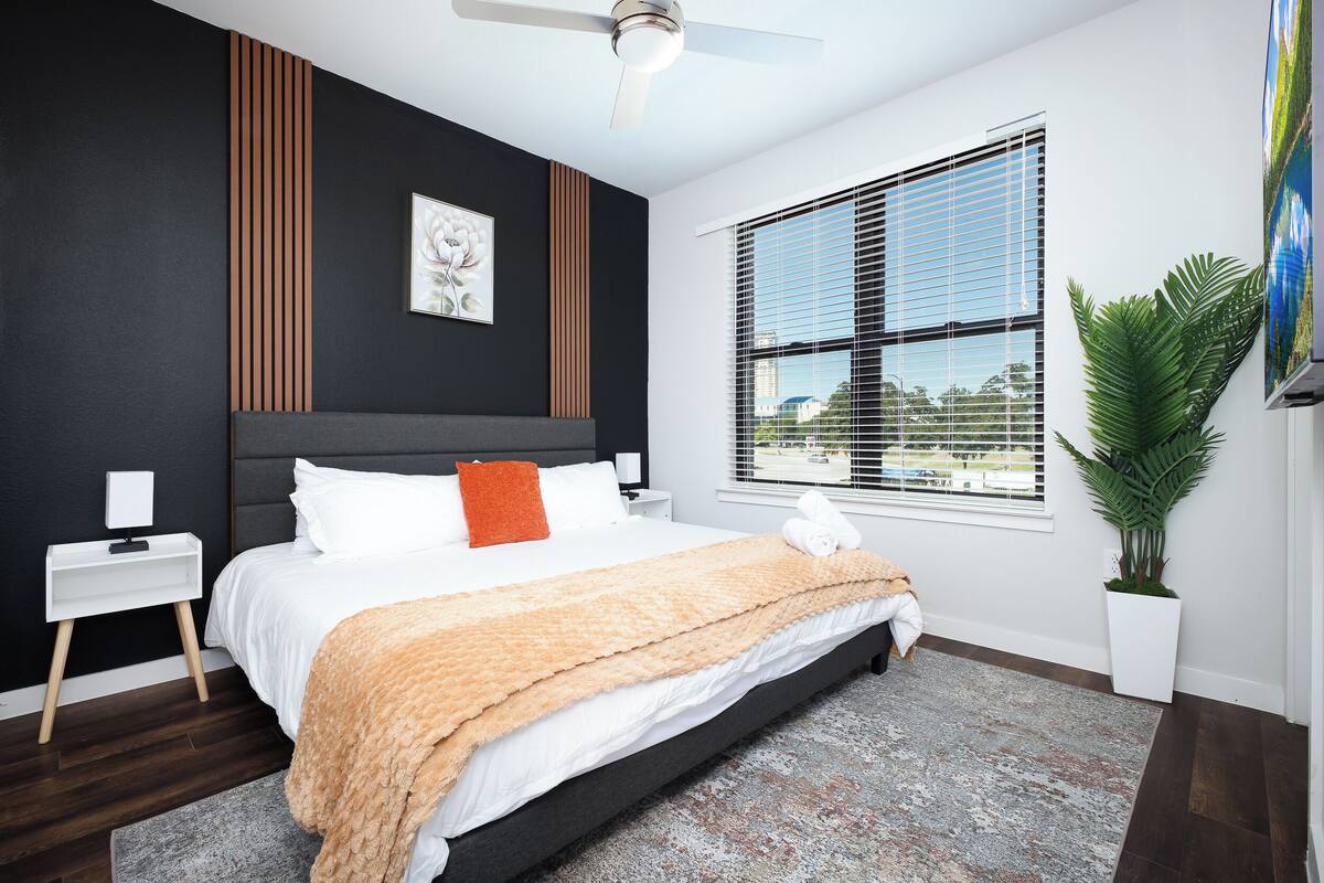 A modern bedroom with a bed, bedside tables, a ceiling fan, a window with blinds, and a potted plant.
