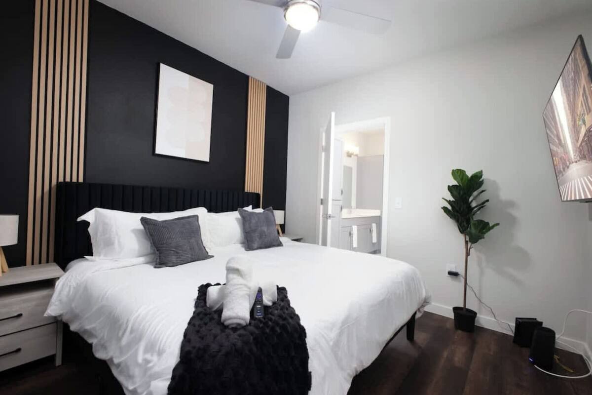 A modern bedroom with a large bed, bedside tables, a ceiling fan, a potted plant, and a view into the bathroom.