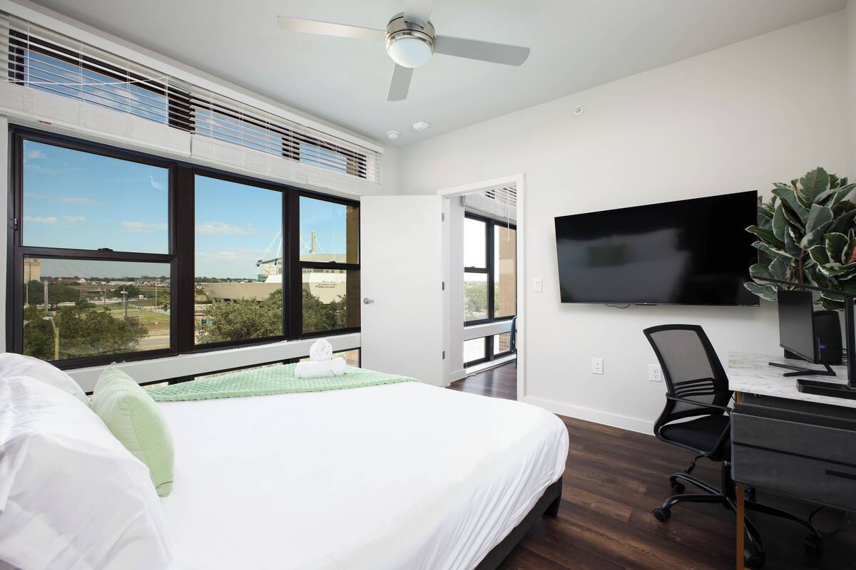 A bedroom with a bed, a desk, a chair, a TV, and large windows with a view.