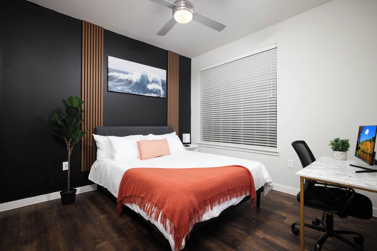A bedroom with a bed, a desk, a chair, a ceiling fan, a window with blinds, and a wall-mounted TV.