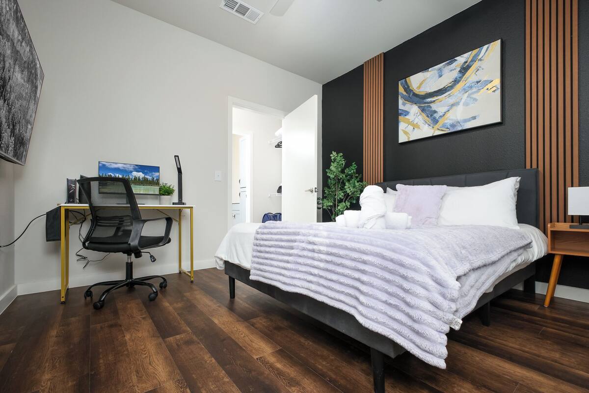 A modern bedroom with a bed, desk, and chair, a painting on the wall, and a door leading to another room.