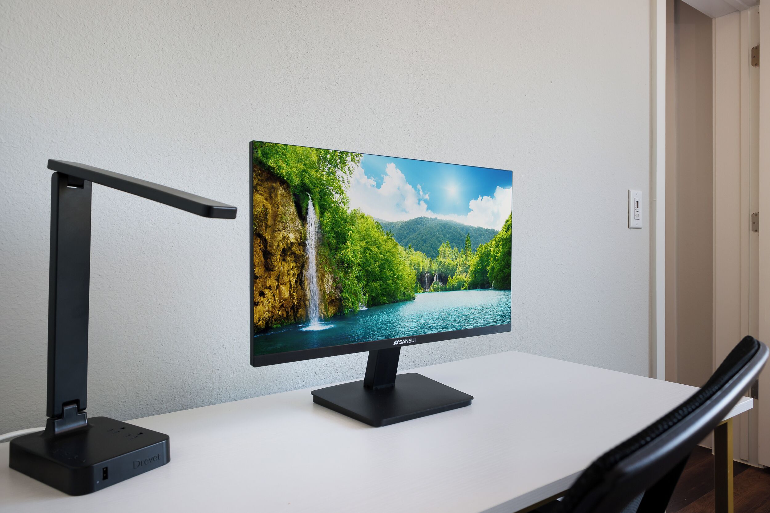 A monitor on a desk with a waterfall and mountains on the screen, a desk lamp, and a chair.