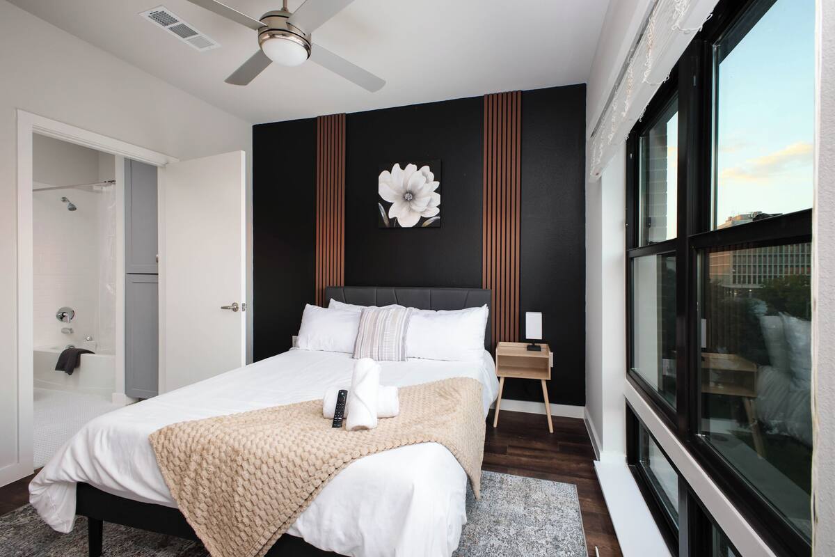A modern hotel room with a large bed, a ceiling fan, a view through a window, and a shower area.