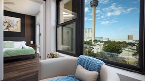 A hotel room with a large window, a bed with green bedding, a white sofa with a blue throw, and a view of a tower and buildings. - Labor by Stylish Stays (San Antonio)