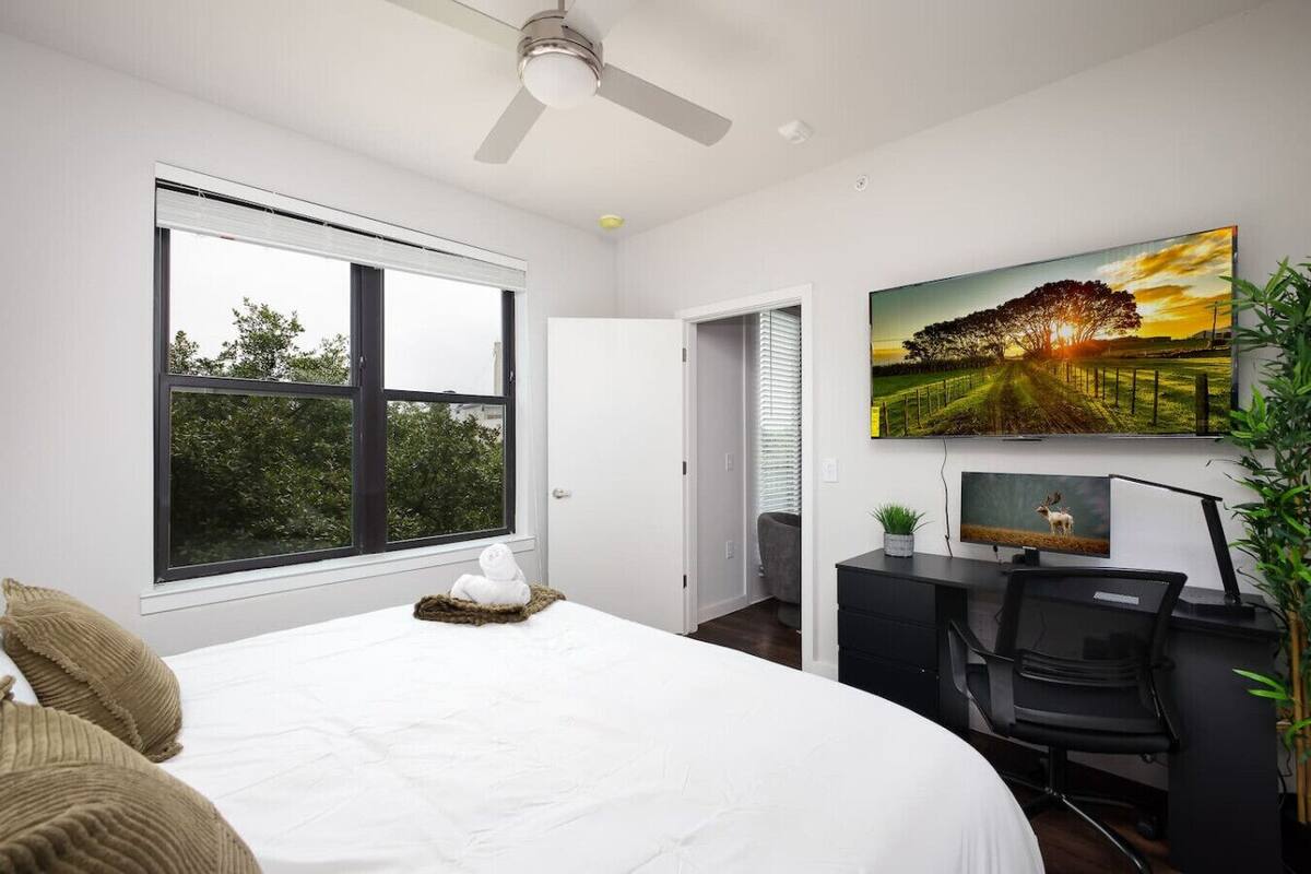 A bedroom with a large bed, a desk, a chair, a television, and a window with a view of trees.