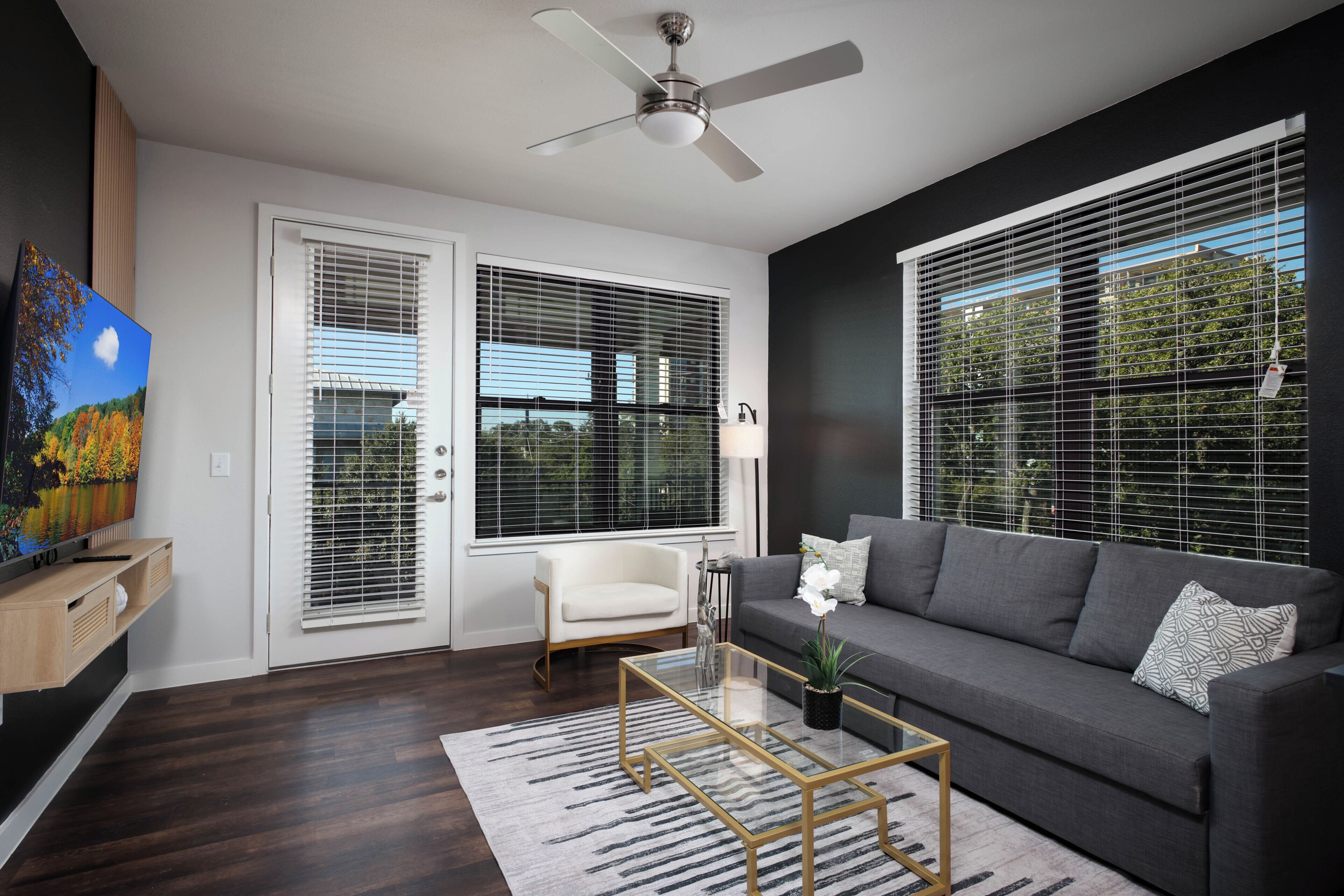 A modern living room with a sofa, a glass coffee table, a ceiling fan, and large windows with blinds.