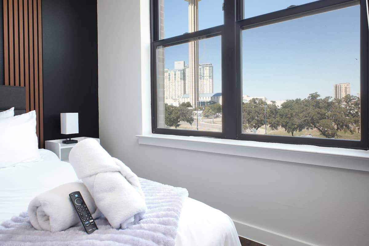 A hotel room with a large window, a bed with white linens, a remote control, and a view of a tower.