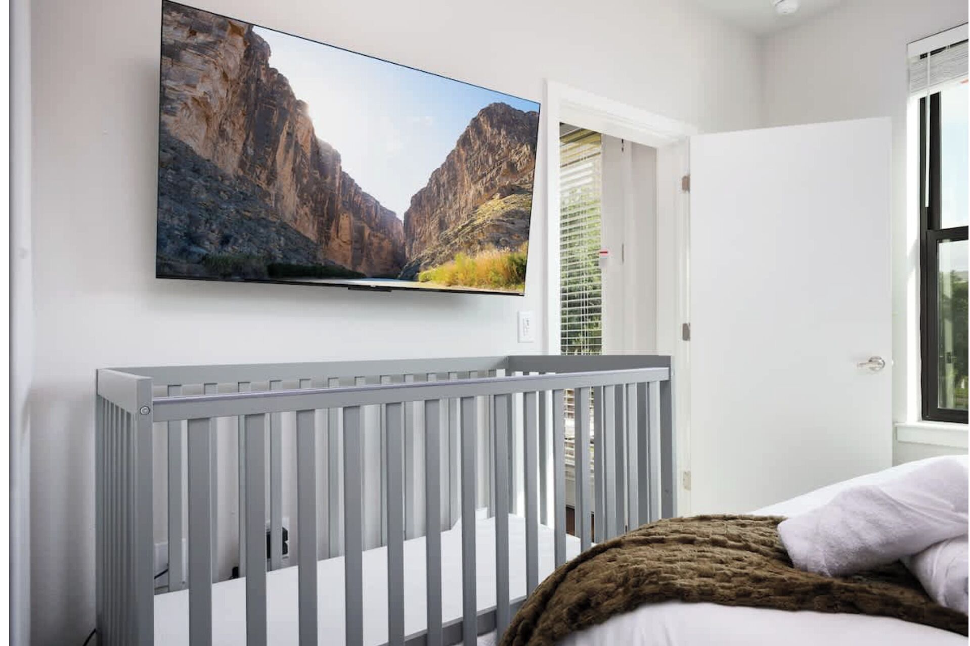 Ein Schlafzimmer mit einem weißen Babybett, einem Wand-Fernseher und einem Fenster mit Jalousien.